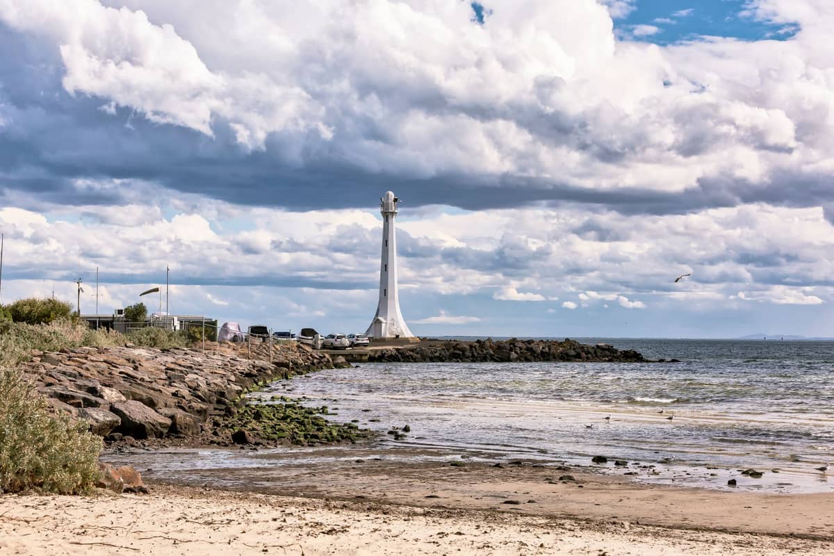 Weitläufiger Blick auf einen Sandstrand und eine felsige Küste mit dem markanten weißen Leuchtturm am Ende des St Kilda Pier, unter einem teils bewölkten Himmel.