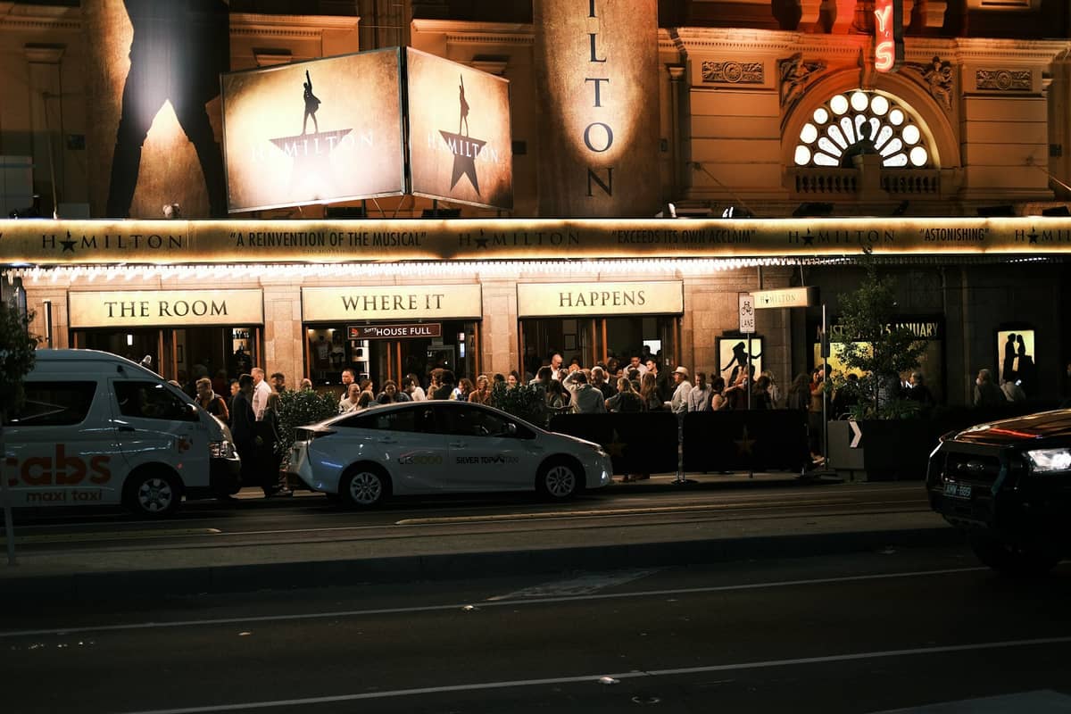 Folla riunita all'ingresso di un teatro di Melbourne di notte, con auto e insegne illuminate visibili.