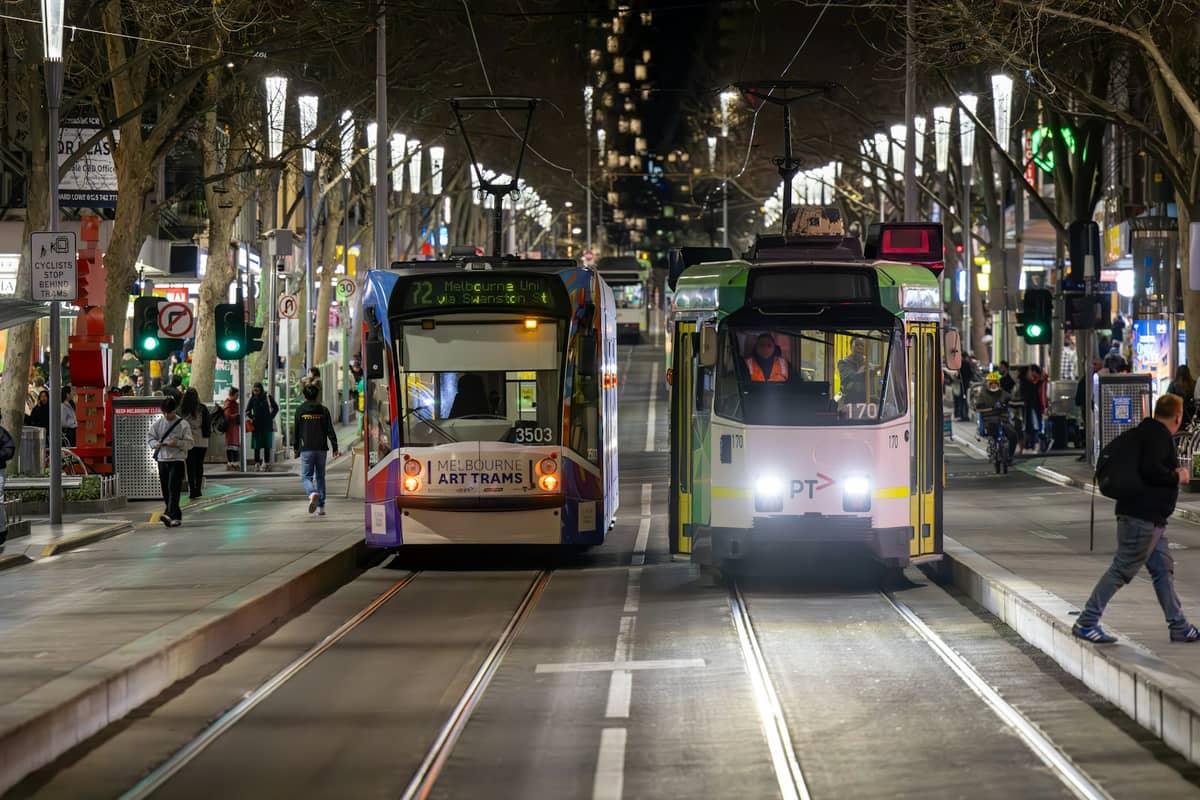 Due tram a una fermata ben illuminata in una strada di Melbourne di notte, con persone che camminano nelle vicinanze e le luci della città visibili.