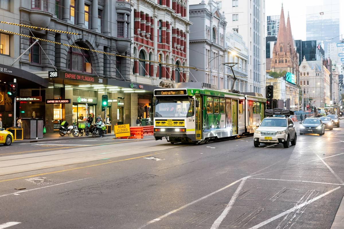 Straßenszene in der Innenstadt von Melbourne in der Abenddämmerung mit einer Straßenbahn, Geschäften und den Türmen der St. Paul's Cathedral im Hintergrund – ein lebendiges multikulturelles Stadtbild.