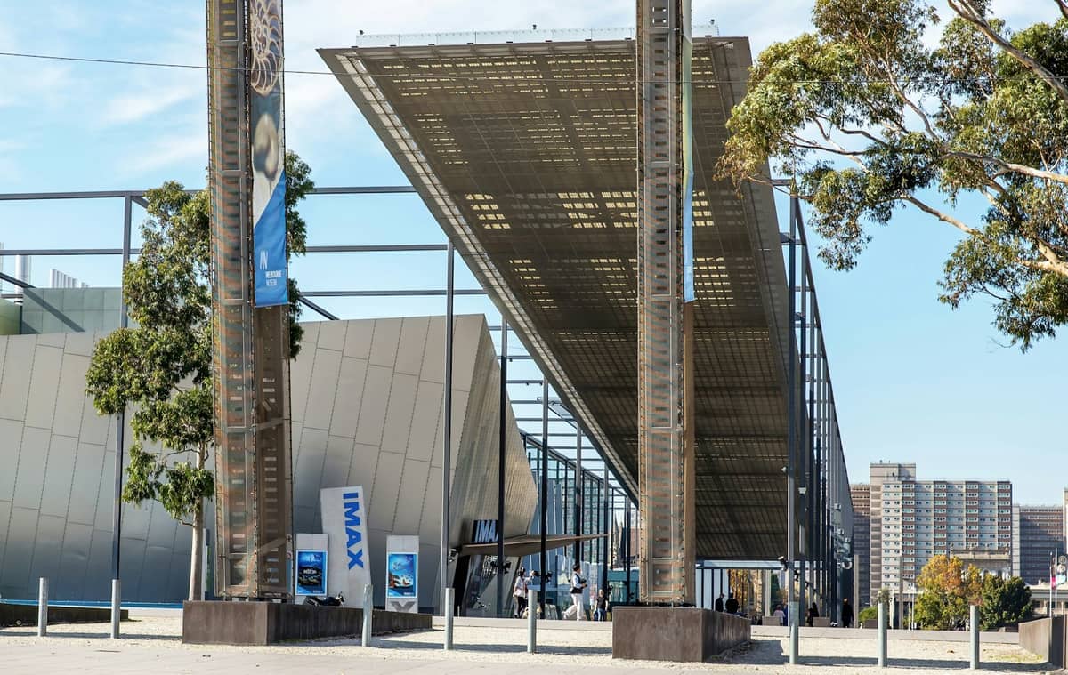 Vista exterior moderna del Melbourne Museum y la entrada del IMAX, con arquitectura contemporánea, altas columnas de metal, árboles y un cielo azul de fondo.