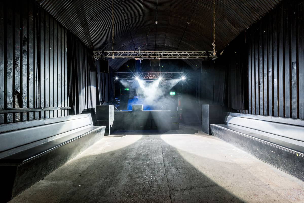 Interior klub yang kosong dengan bangku di sepanjang dinding, booth DJ, lampu panggung, dan dekorasi industrial.