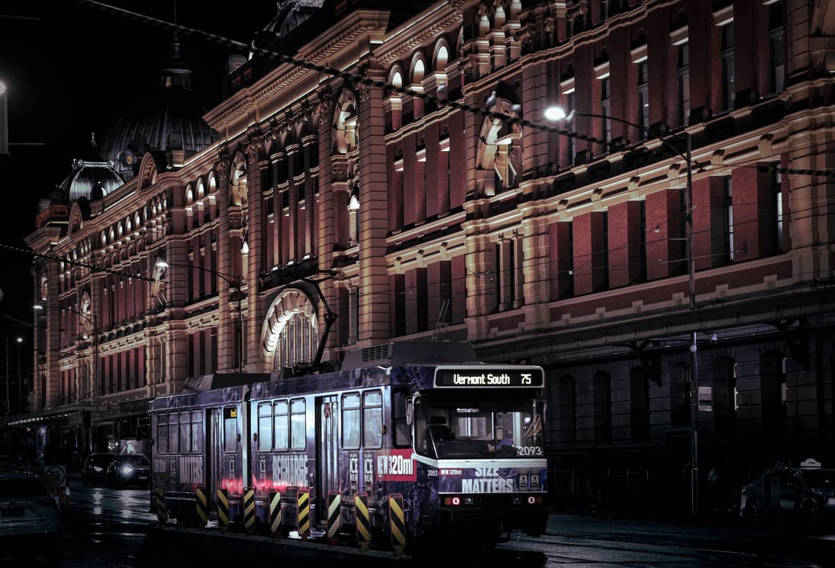Pemandangan malam trem Melbourne di depan Flinders Street Station, dengan lampu-lampu dan bangunan ikonik di latar belakang.