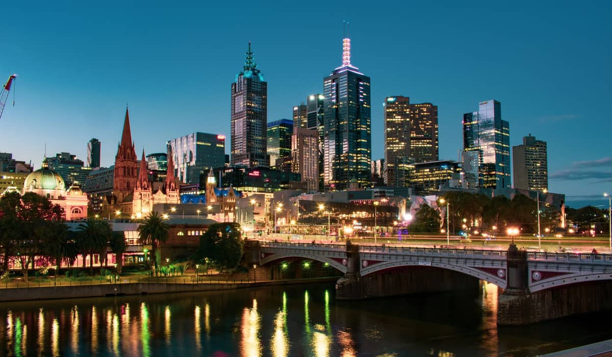 Vista nocturna del horizonte de Melbourne con rascacielos iluminados, la catedral de San Pablo y un puente reflejados en el río Yarra, capturando el ambiente vibrante de la vida nocturna.