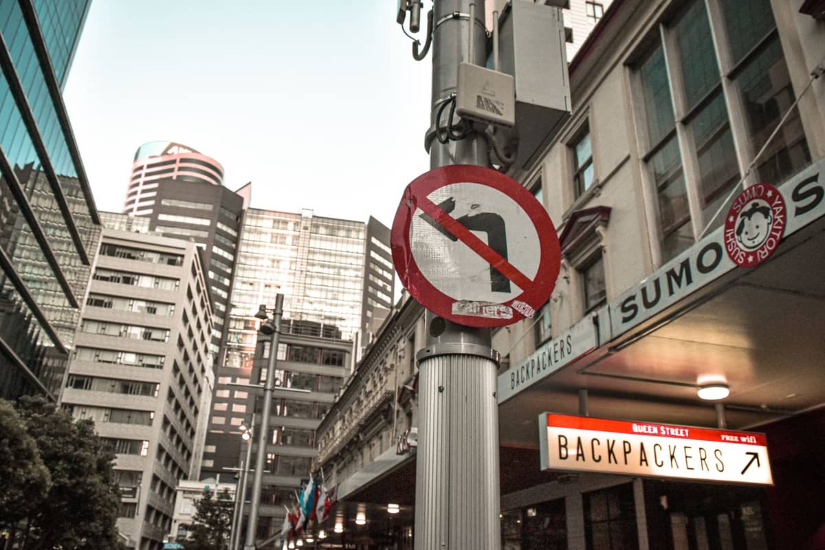 Vue au niveau de la rue d'un pâté de maisons avec une enseigne lumineuse intitulée 'Backpackers' à côté de grands immeubles modernes, suggérant un hébergement en auberge de jeunesse abordable en centre-ville.