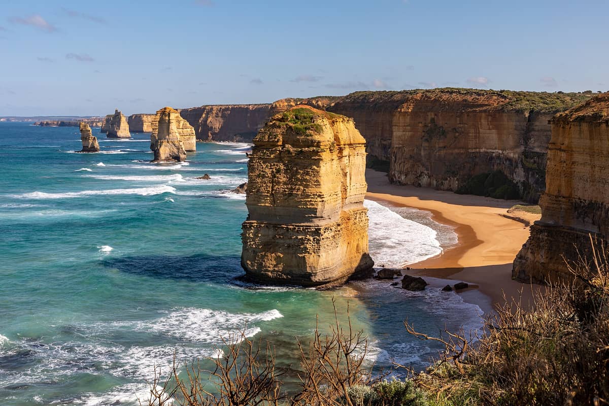 Dramatische Küstenansicht der Twelve Apostles Felsformationen, goldener Klippen und türkisfarbener Wellen an Australiens Great Ocean Road unter einem sonnigen Himmel.