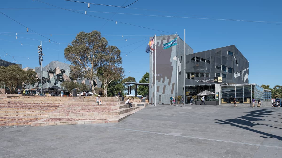 Federation Square in Melbourne mit seiner markanten modernen Architektur und Treppen, entspannenden Menschen und klarem blauem Himmel.