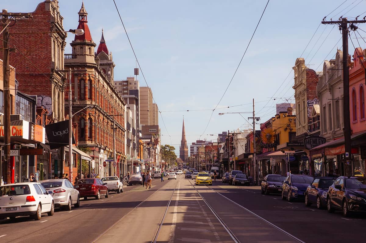 Straßenansicht der Brunswick Street in Fitzroy mit historischen Gebäuden, Straßenbahnschienen, Geschäften, Autos und Fußgängern an einem sonnigen Tag.