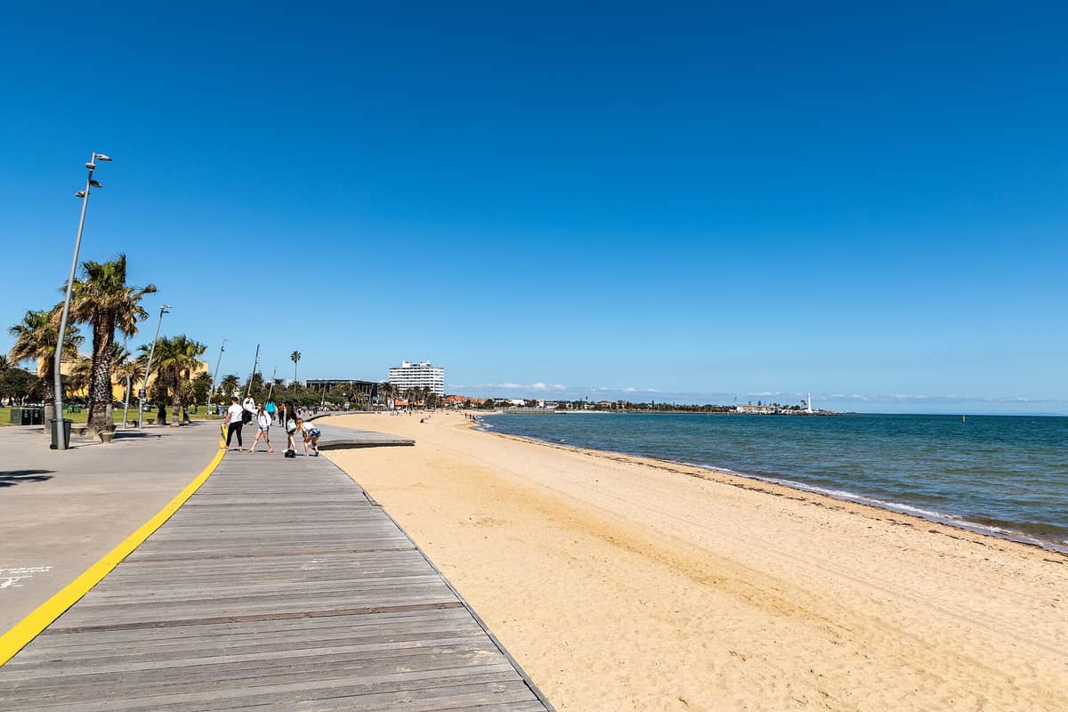 Weitwinkelansicht der Strandpromenade von St Kilda Beach mit Menschen, Palmen, Sandstrand und ruhigem blauem Wasser an einem sonnigen Tag.