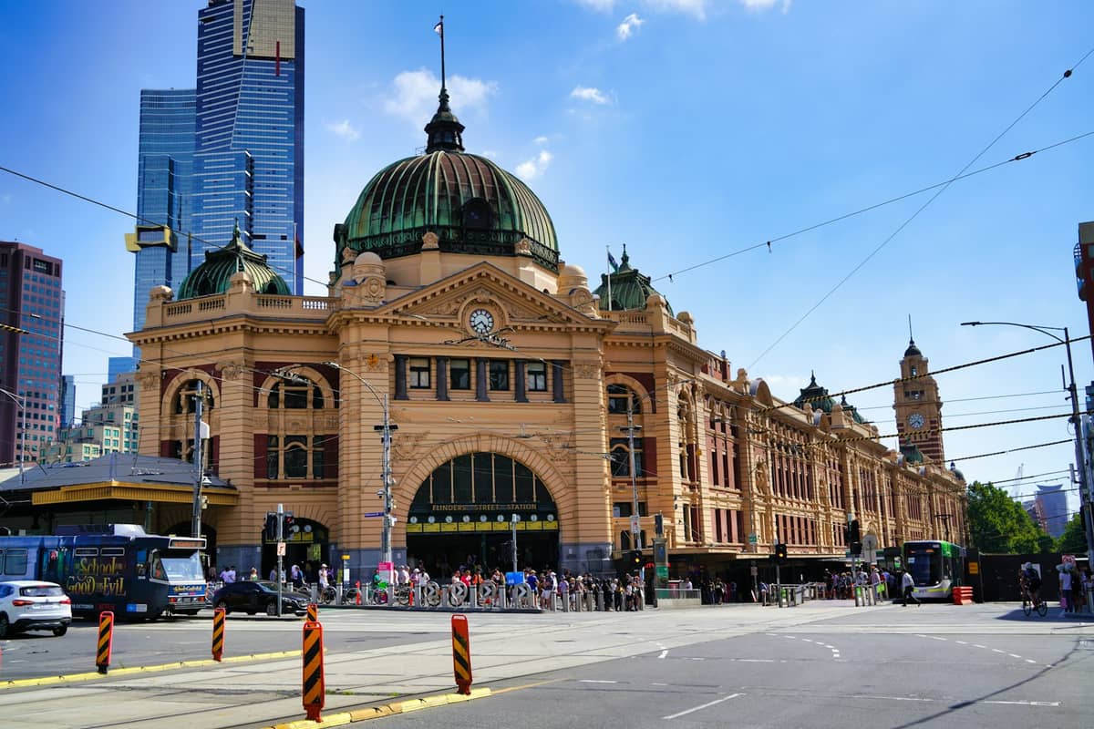 Vista amplia de la Estación Flinders Street en Melbourne con su icónica cúpula, torre y rascacielos circundantes bajo un cielo azul.