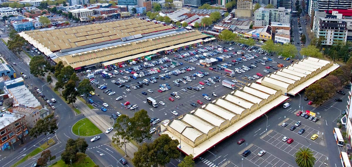 Luftaufnahme der Gebäude des Queen Victoria Market in Melbourne und des angrenzenden großen Parkplatzes, umgeben von Stadtstraßen und städtischen Gebäuden.