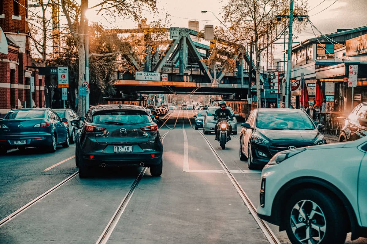 Straßenansicht der Brunswick Street in Fitzroy, Melbourne, gesäumt von Geschäften, Cafés, Autos und einer Eisenbahnbrücke in der Ferne bei Sonnenuntergang.