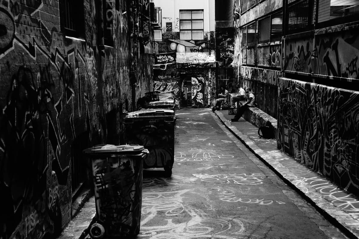 Foto em preto e branco de uma viela de Melbourne coberta de grafites e arte de rua, com uma pessoa sentada num banco.