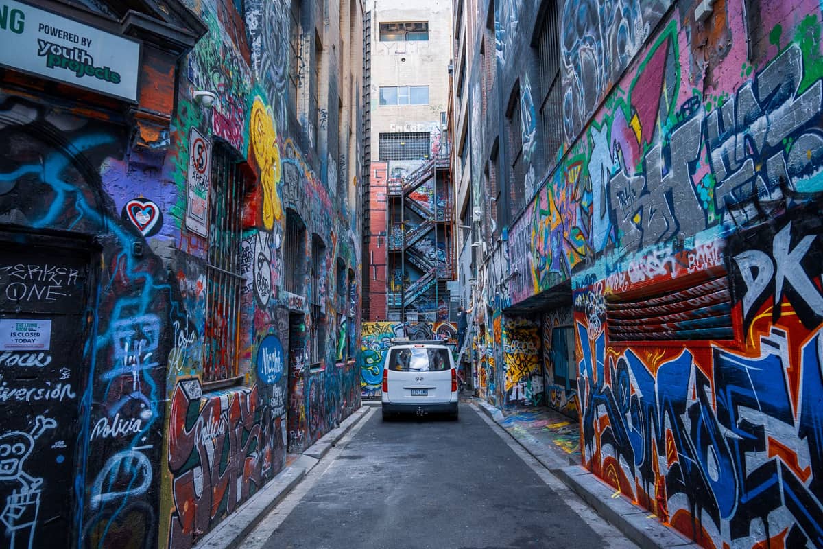 Uma estreita viela de Melbourne coberta do chão ao teto com arte de rua colorida e grafite, com uma van branca estacionada no centro.