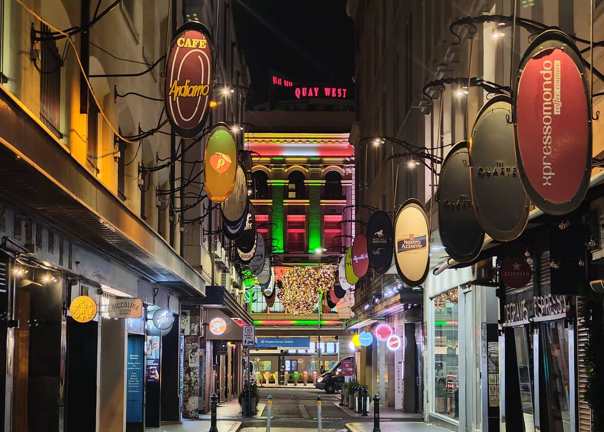 Lebhafte Gasse in Melbourne bei Nacht mit beleuchteten Schaufenstern, bunten Schildern und einem gut beleuchteten Gebäude im Hintergrund, einladend für Fußgänger.