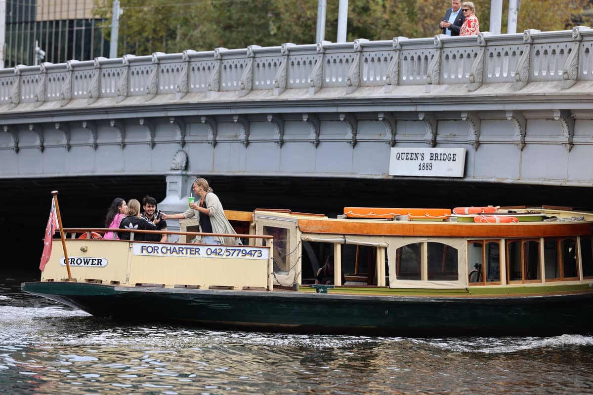 Familias y niños a bordo de un barco charter bajo el Queen's Bridge sobre el río Yarra, en el centro de Melbourne, con el paisaje urbano de fondo.