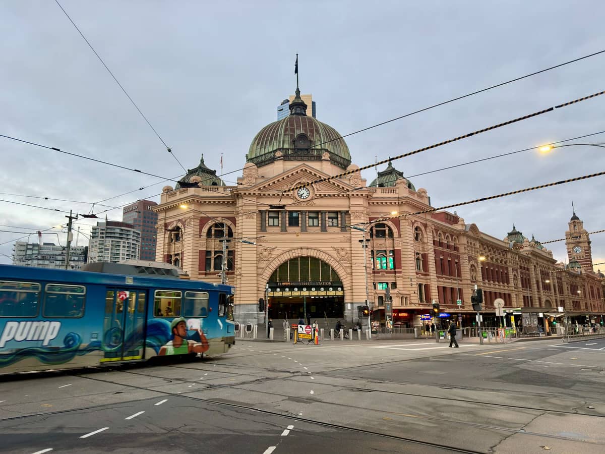 Eine Straßenbahn fährt an einem bewölkten Tag vor dem Flinders Street Bahnhof in Melbourne mit sichtbaren Straßen- und Oberleitungsdrähten vorbei.