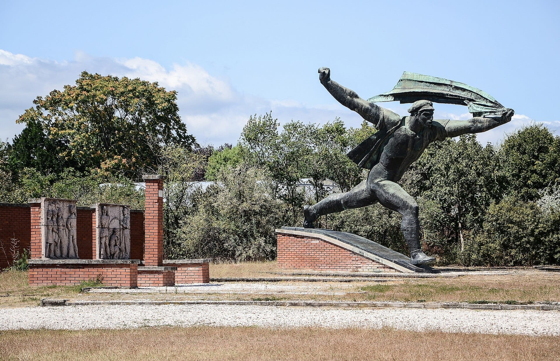 Große Bronzestatue eines Mannes mit einer Fahne im Memento Park Budapest, umgeben von Bäumen und roten Backsteinmonumenten unter einem klaren Himmel.