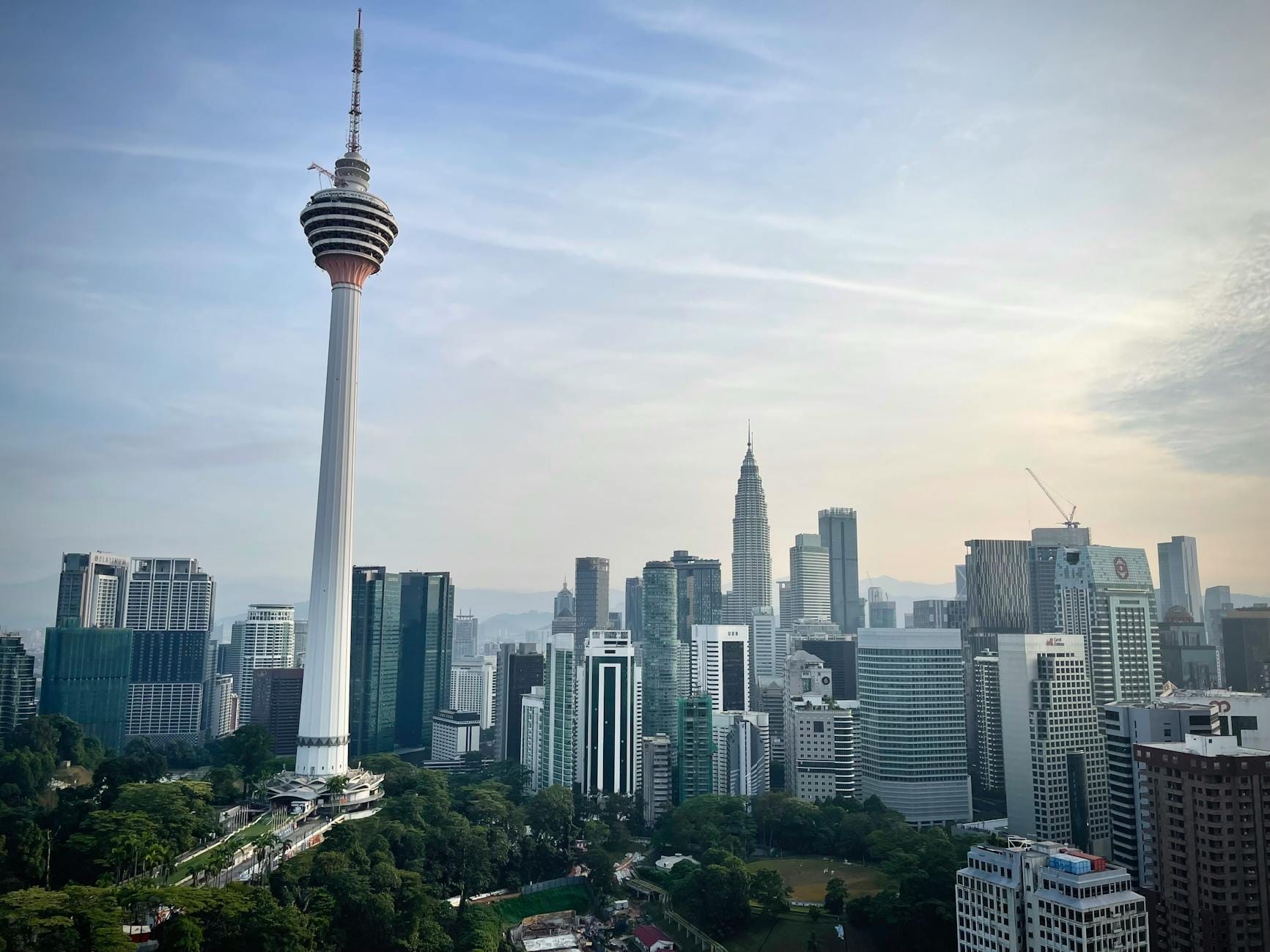 Menara KL Tower stands tall above the Kuala Lumpur skyline, surrounded by modern skyscrapers and lush greenery under a partly cloudy sky.