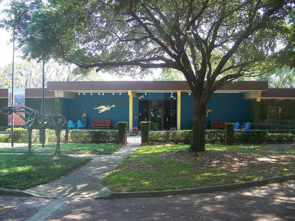Front exterior of the Mennello Museum of American Art with its blue facade, sculpture garden, large tree, and main entrance visible on a sunny day.