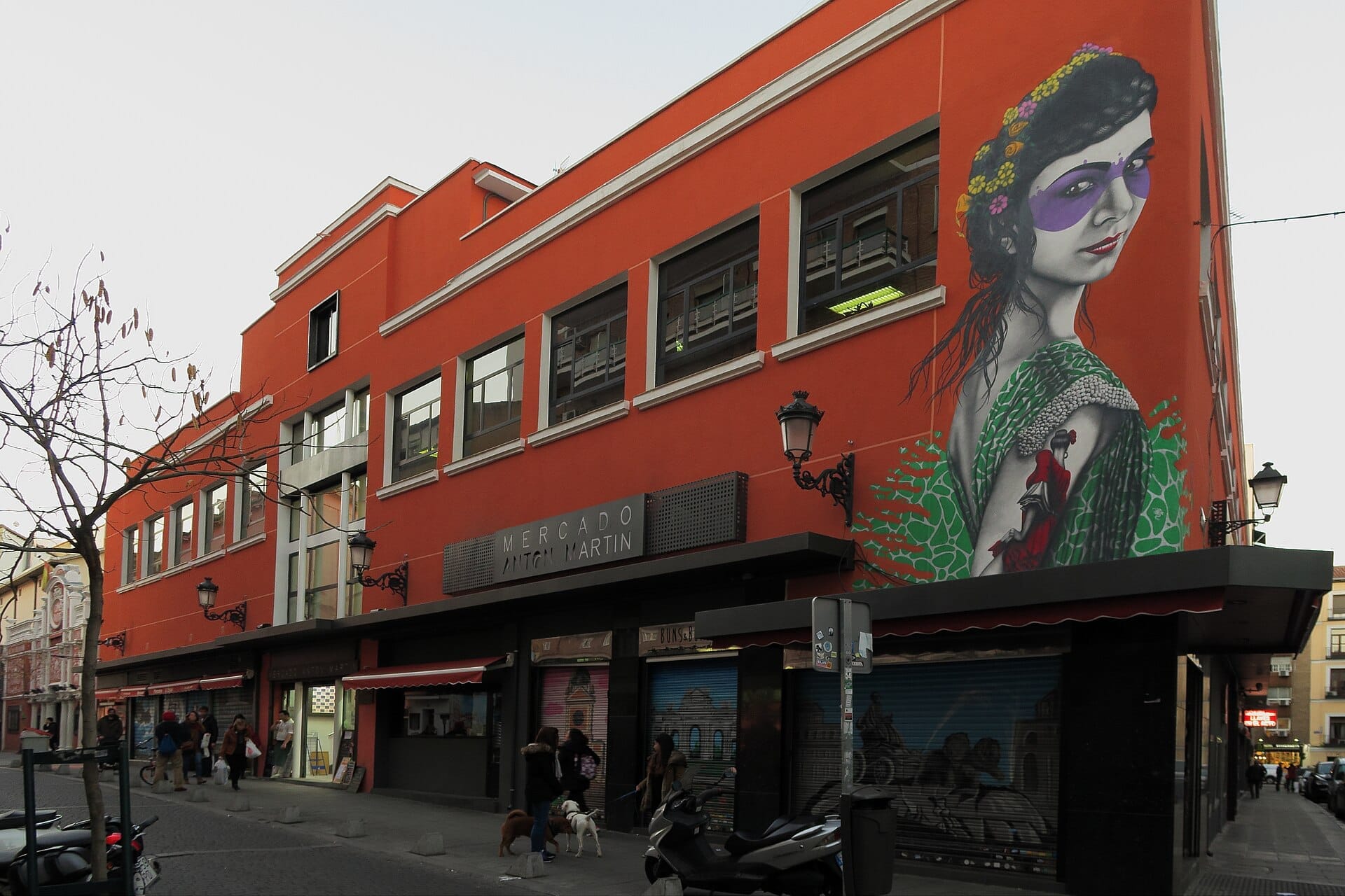 Esterno del Mercado de Antón Martín con pareti arancioni brillanti, grandi finestre, un murale di una donna e persone che portano a spasso i cani sul marciapiede.