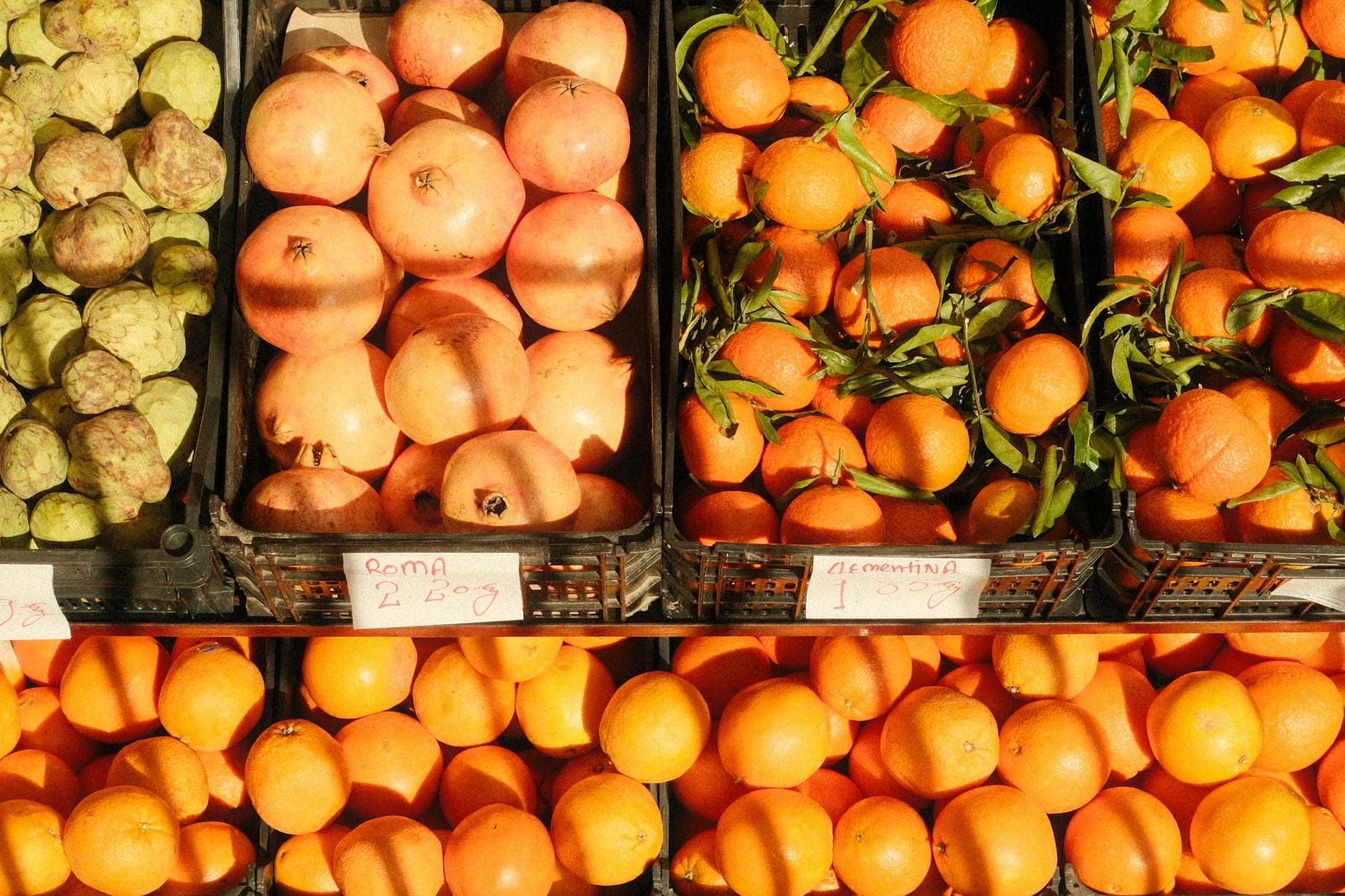 Primo piano di prodotti freschi in cassette di mercato, tra cui melograni, arance e frutti verdi, illuminati in modo vivido e invitante, che catturano l'atmosfera autentica del mercato locale.
