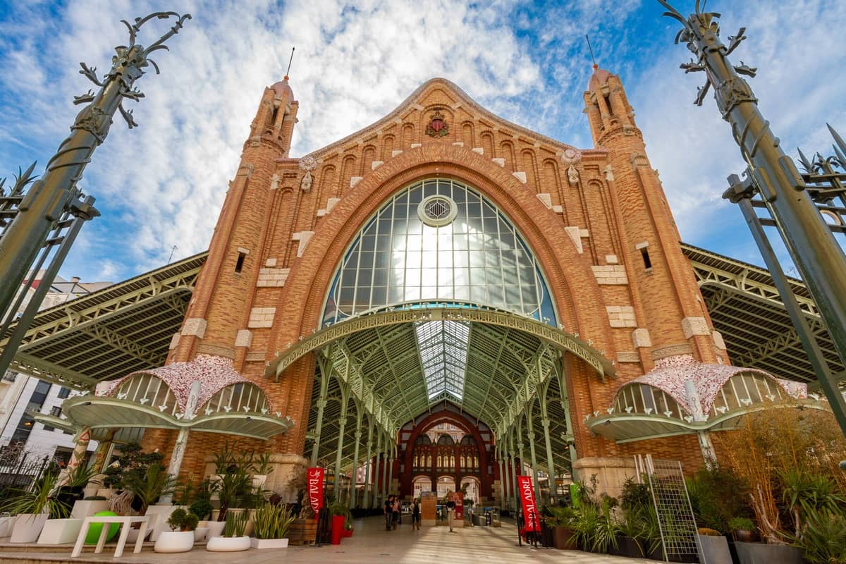 Vista grandangolare della facciata modernista in mattoni del Mercado de Colón a Valencia, con l'imponente ingresso ad arco in vetro, le torri decorative e le persone che entrano nel mercato.
