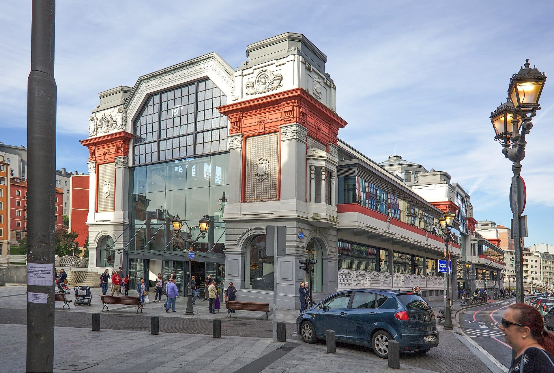 Das Gebäude des Mercado de la Ribera in Bilbao mit seinem großen Bogeneingang, roter und weißer Fassade und Menschen, die draußen spazieren.