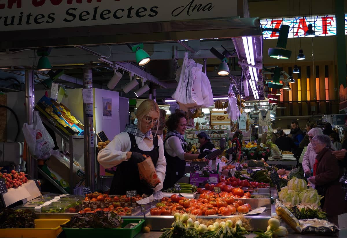 Un banco del mercato pieno di frutta e verdura all'interno del Mercado de Ruzafa, con venditori e clienti del quartiere, pomodori e ortaggi esposti sotto la luce al neon e una parte dell'insegna con vetrate colorate del mercato visibile sullo sfondo.