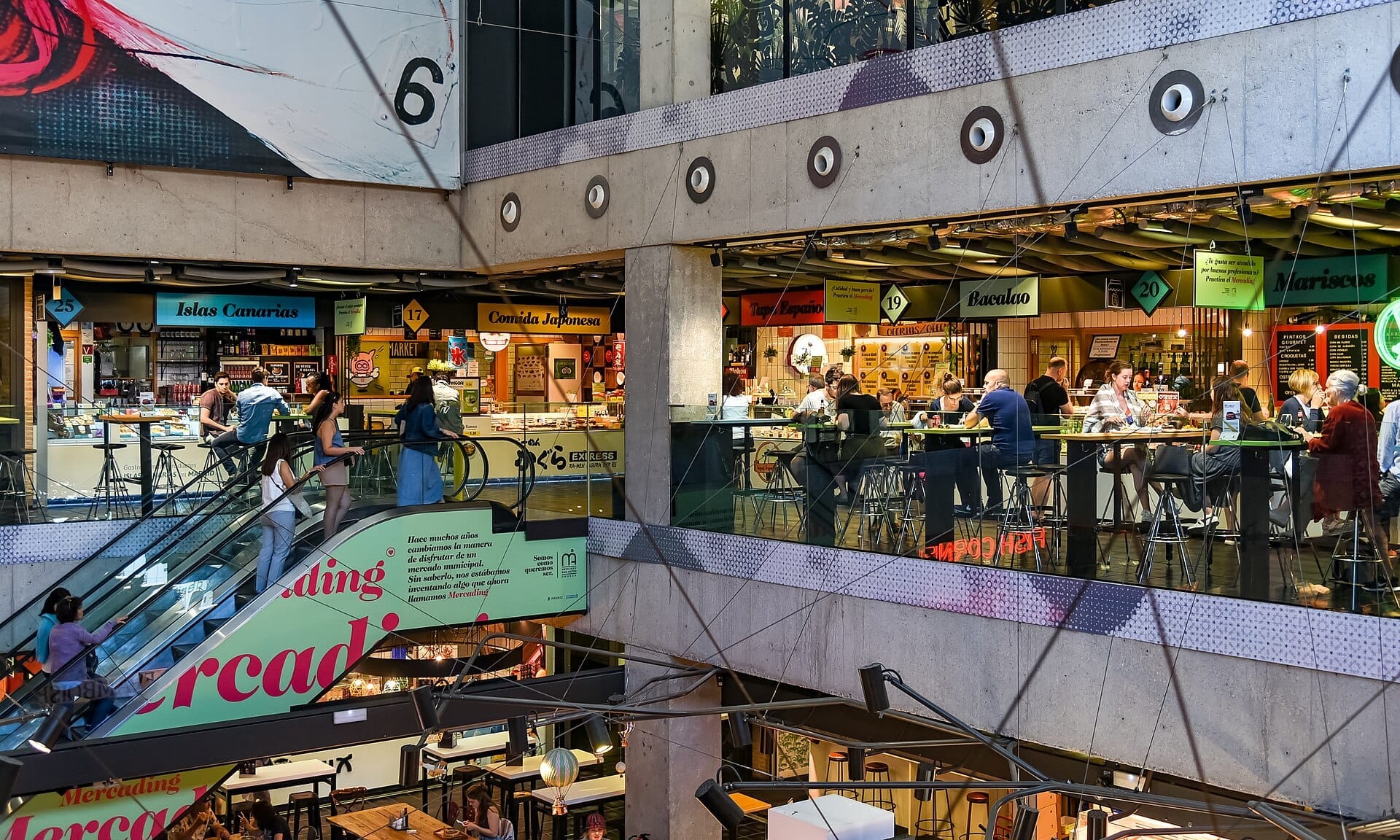 Vista interna del Mercado de San Antón con diversi banchi di cibo, scale mobili e persone che cenano a tavoli moderni su più piani.