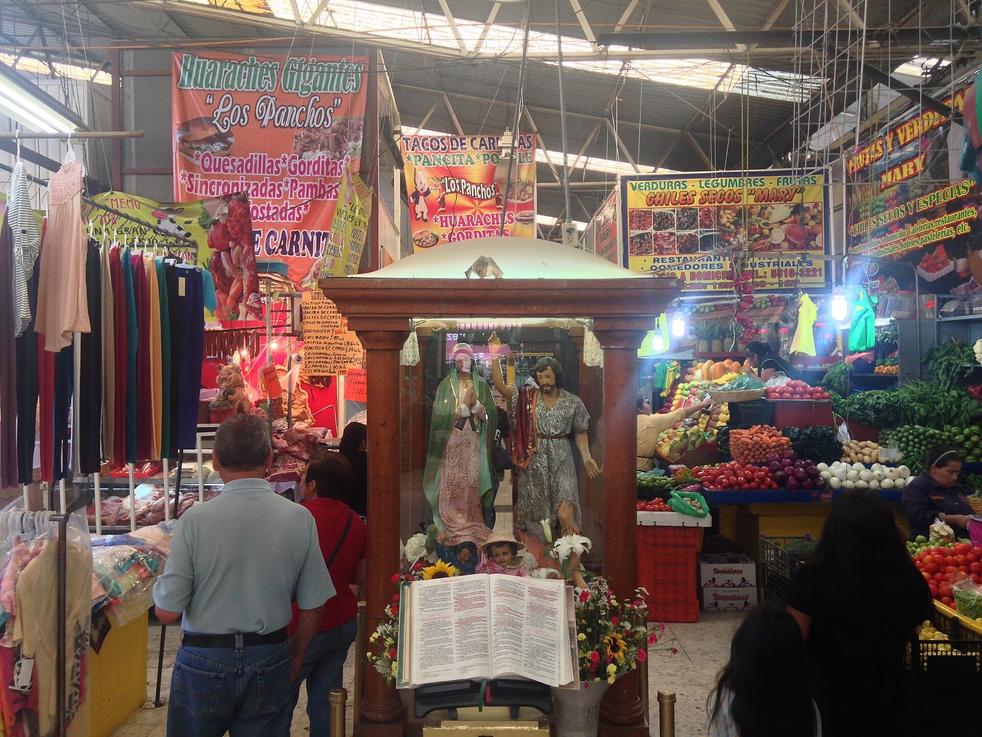 L'interno vivace del Mercado de San Juan con bancarelle colorate di prodotti, statue religiose, abiti appesi e acquirenti sotto luci fluorescenti.