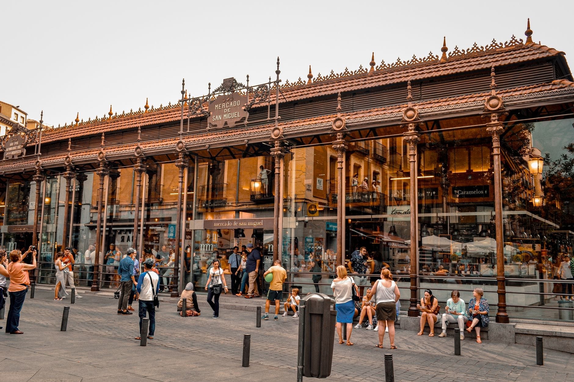 Vista d'insieme della facciata ornata in vetro e ferro del Mercado de San Miguel con persone radunate all'esterno, che cattura l'atmosfera vivace nel cuore di Madrid.