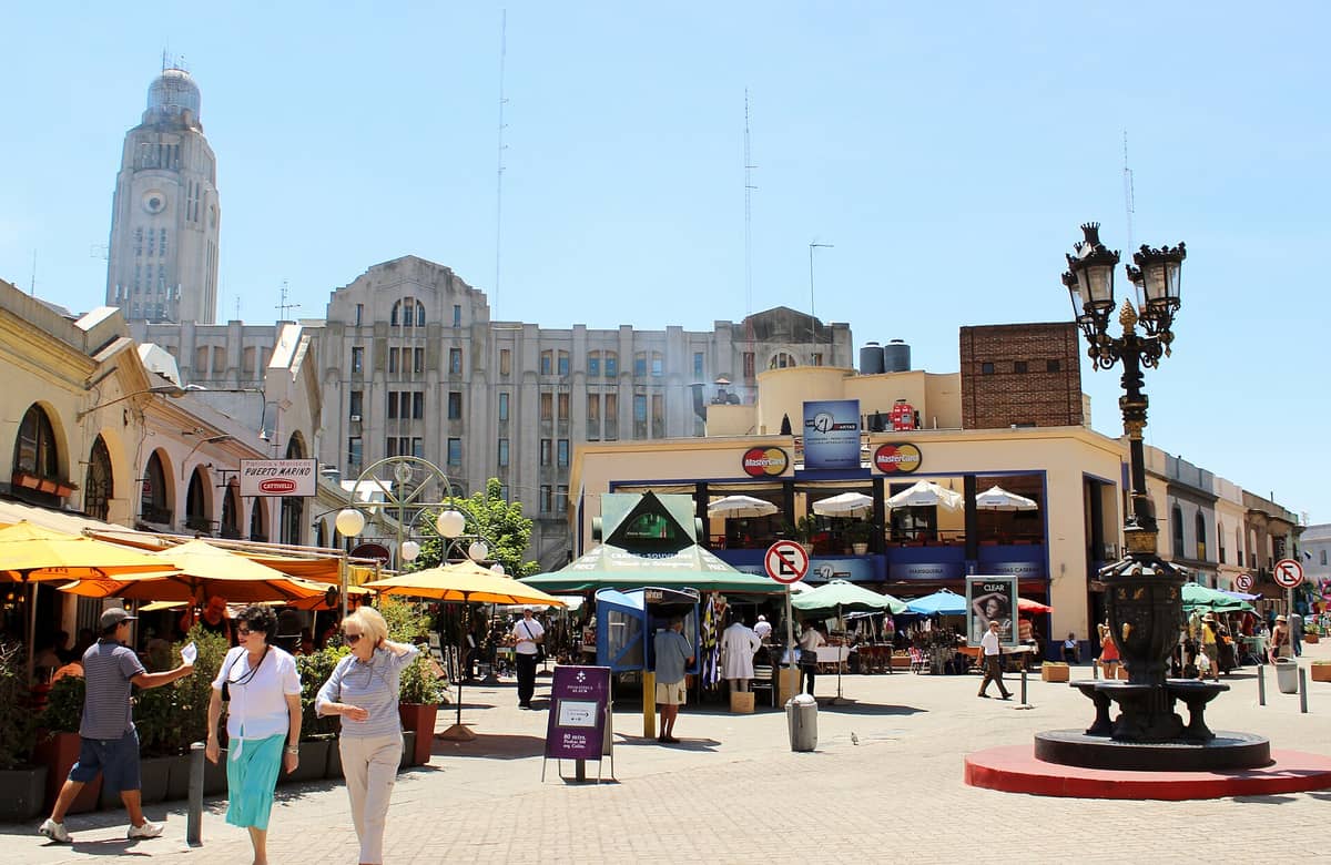 ลานหน้าตลาด Mercado del Puerto ในมอนเตวิเดโอ คนเดินไปมา ร่มร้านค้า และอาคารท่าเรืออาร์ตเดโคอยู่ด้านหลัง