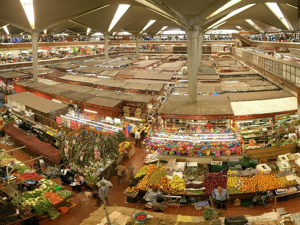 Vista interna del Mercado San Juan de Dios con file di bancarelle, prodotti colorati e acquirenti sotto un grande tetto moderno.