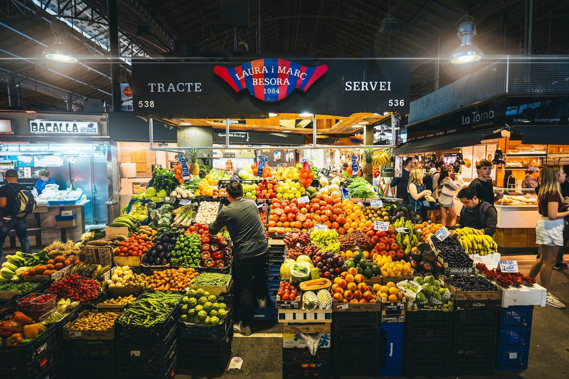 A vibrant fruit and vegetable stall in the heart of La Boqueria market with shoppers browsing and colorful produce displayed under the market roof.