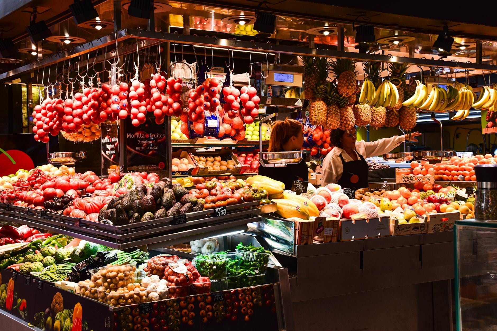 Colorato banco di frutta e verdura al Mercat de Santa Catalina, con pomodori, banane, ananas e acquirenti sotto una calda illuminazione interna.