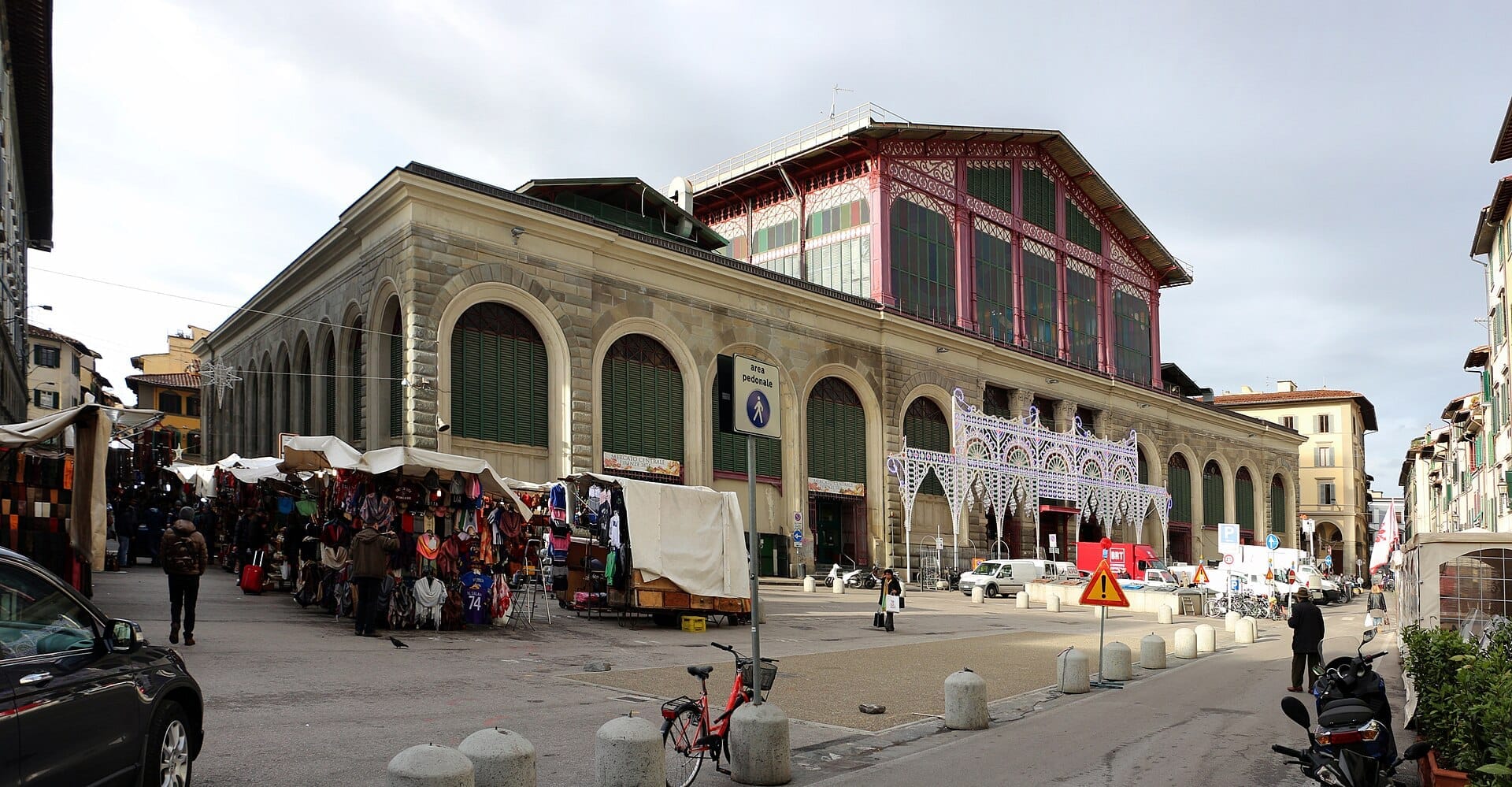 วิวด้านนอกของ Mercato Centrale Florence ที่มีสถาปัตยกรรมหินและกระจกอันทรงประวัติศาสตร์ พ่อค้าแม่ขายริมถนน คนเดินเท้า และท้องฟ้าที่มีเมฆมาก