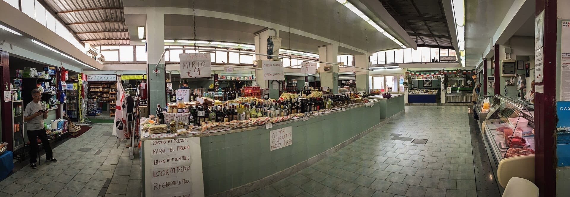Weitwinkelansicht des Inneren des Mercato di San Benedetto mit verschiedenen Lebensmittelständen, Marktverkäufern, Regalen mit Flaschenwaren und natürlichem Licht durch große Fenster.