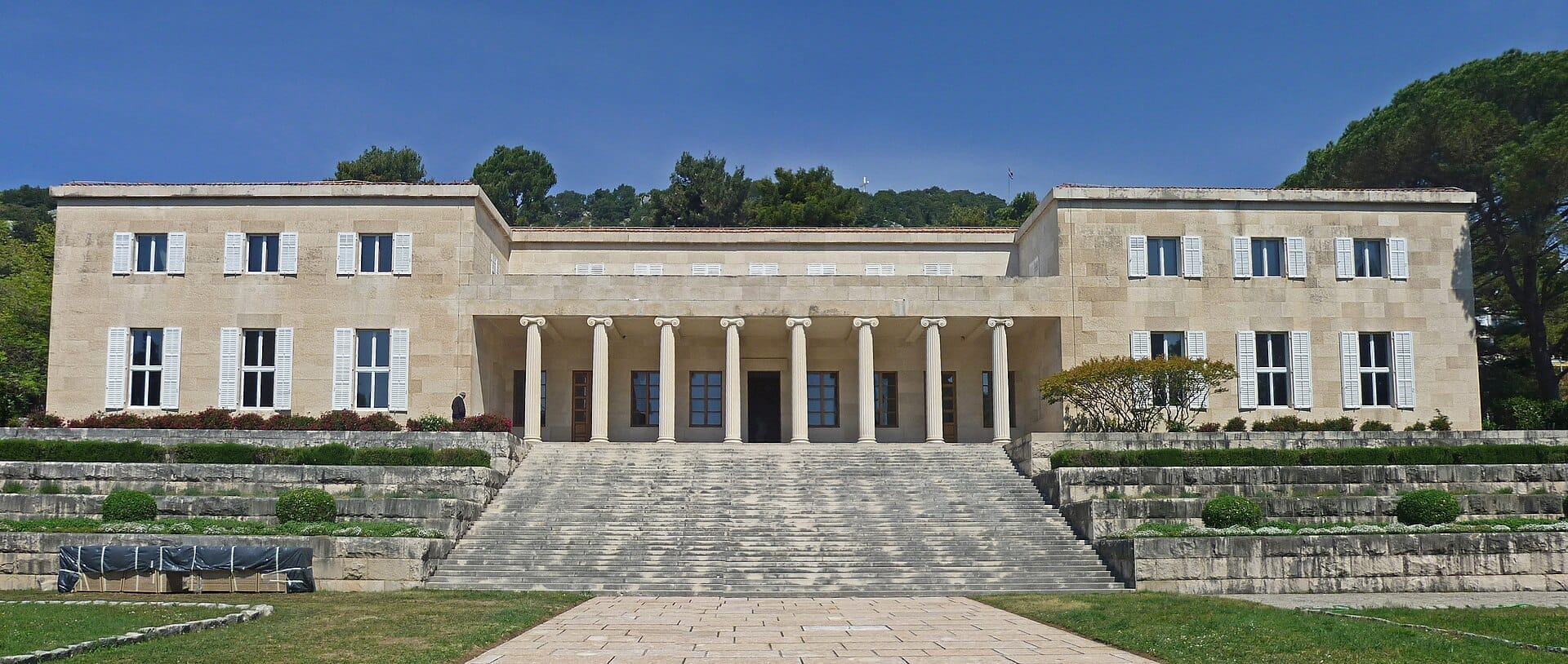 มุมมองด้านหน้าของ Meštrović Gallery ที่แสดงให้เห็นด้านหน้าแบบนีโอคลาสสิกที่สมมาตร เสาสูง บันไดหิน และพุ่มไม้ที่ตัดแต่งแล้วภายใต้ท้องฟ้าสีฟ้าใส
