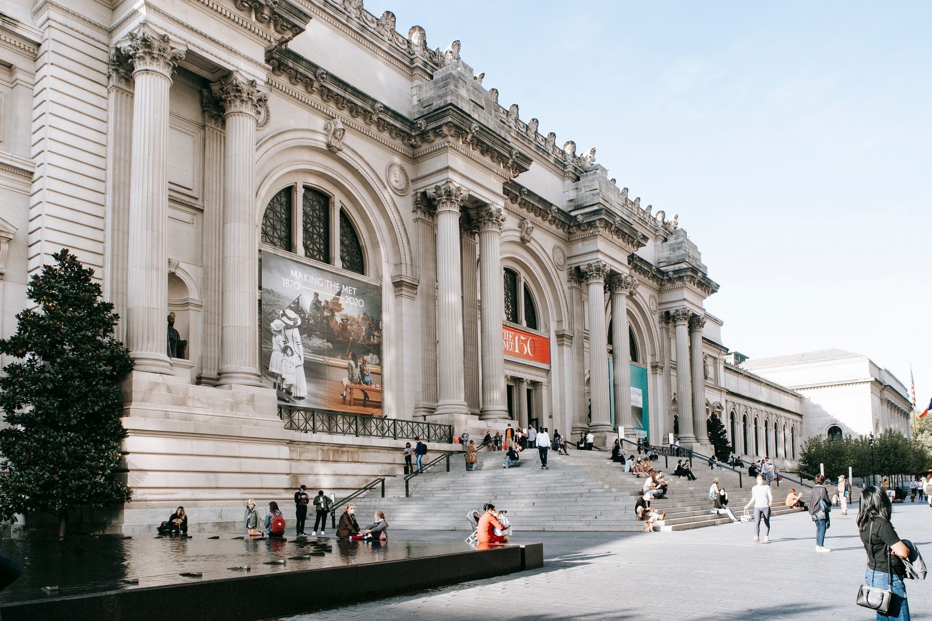 Vista ampia della maestosa facciata del Metropolitan Museum of Art con le iconiche colonne, la scalinata e i visitatori seduti e in cammino fuori dal museo in una giornata di sole.