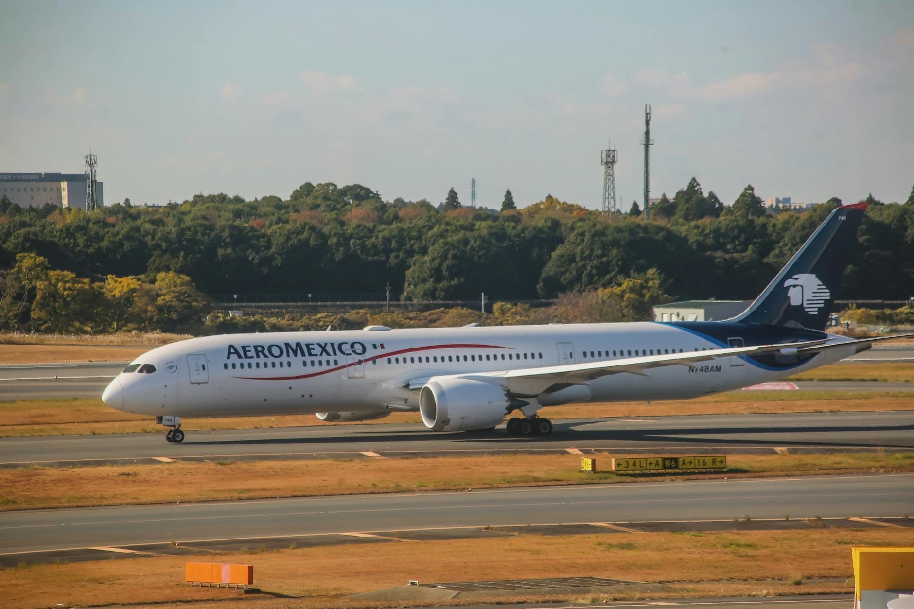 Avião da Aeromexico taxiando na pista de um aeroporto com árvores e infraestrutura ao fundo sob um céu limpo.