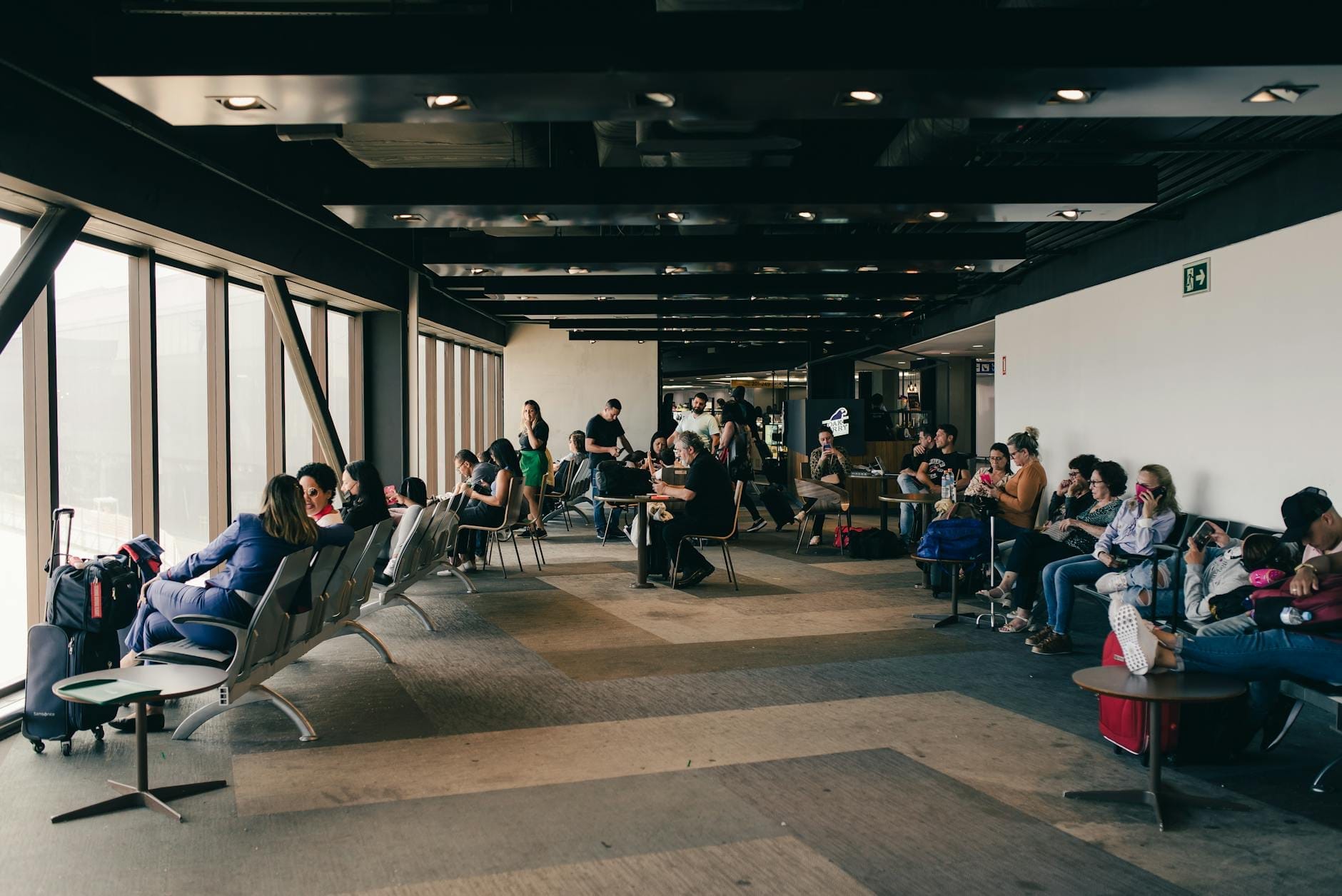 Viajantes sentados e esperando em uma moderna área de sala de embarque de aeroporto com grandes janelas e assentos visíveis.