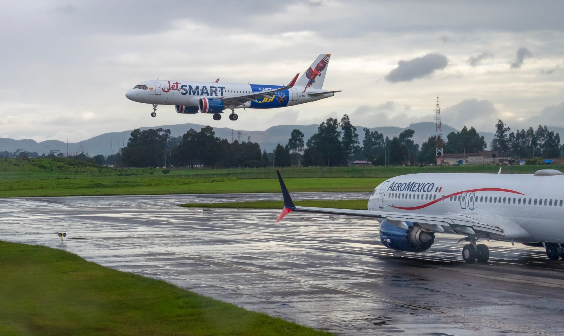 Dois aviões comerciais, um da JetSMART pousando e um da Aeroméxico taxiando, em uma pista molhada com vegetação verde e céus nublados.