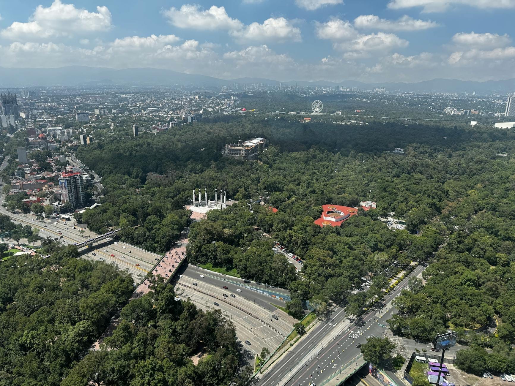 Vista aérea da Cidade do México com o Chapultepec Park, edifícios da cidade e montanhas ao fundo sob um céu parcialmente nublado
