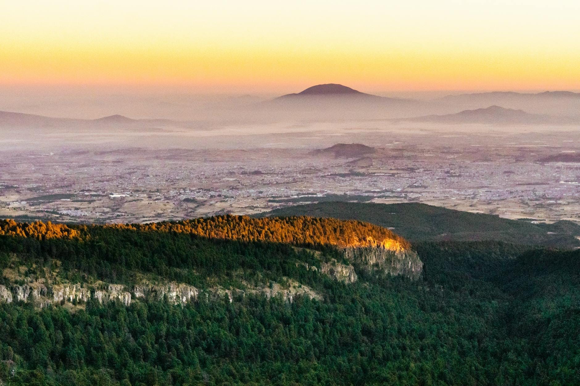 Vista panorâmica de cima mostrando a Cidade do México se espalhando por um amplo vale com montanhas distantes e cristas arborizadas ao amanhecer.
