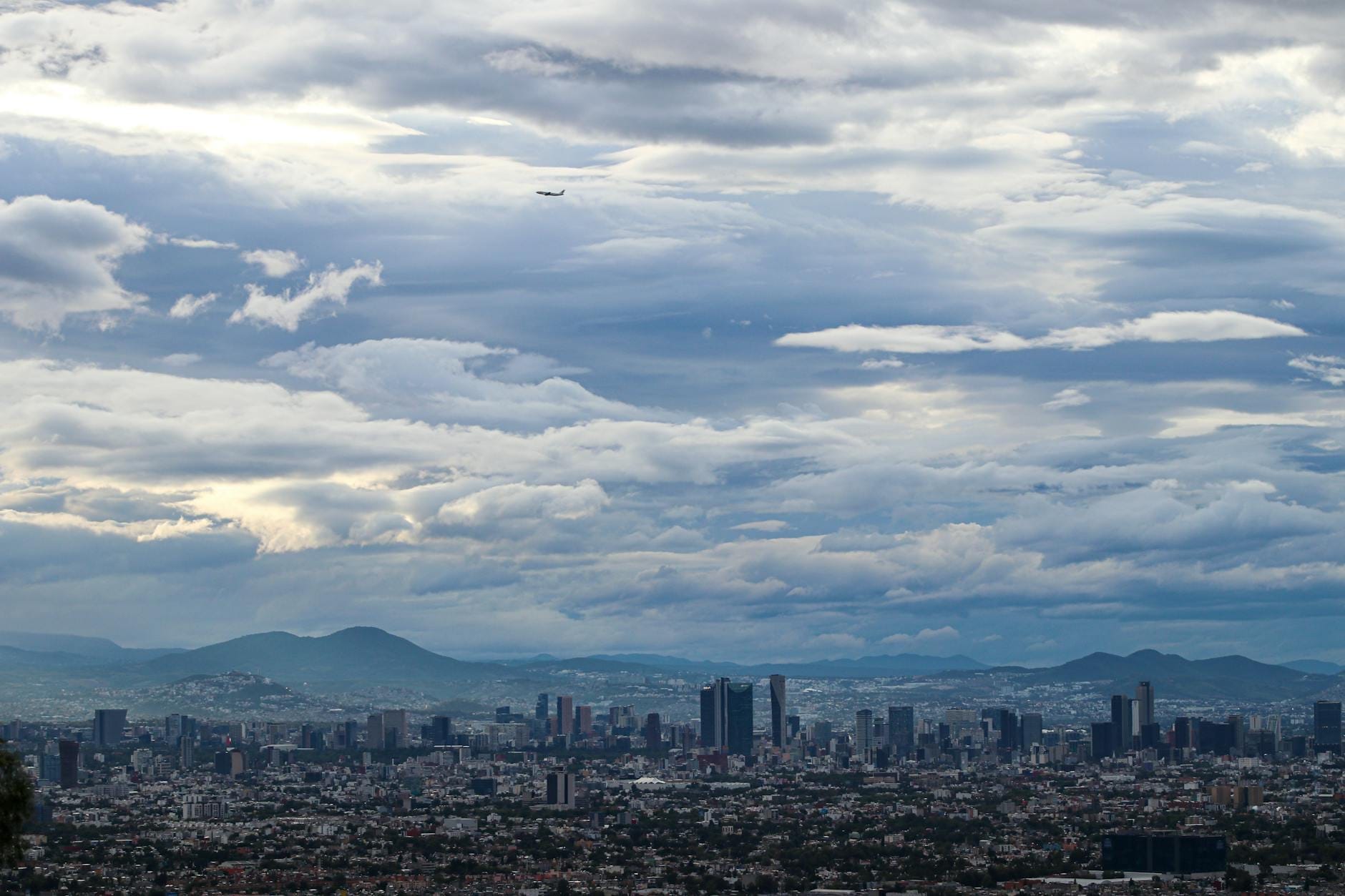 Vista aérea ampla da Cidade do México com edifícios densamente agrupados, montanhas ao fundo e um avião voando sob um céu dramático.