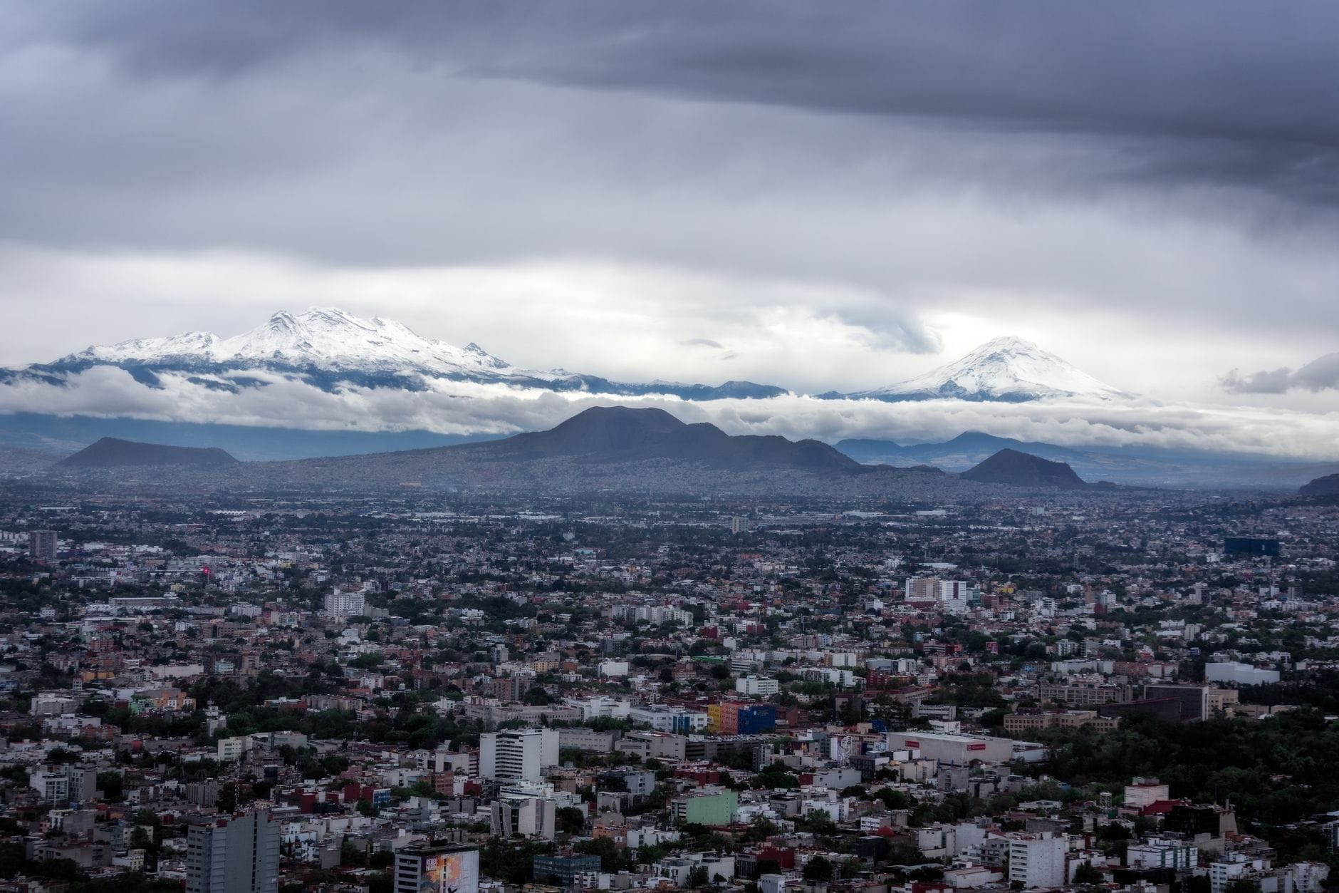 Vista panorâmica da Cidade do México com nuvens dramáticas e vulcões cobertos de neve ao fundo, mostrando a cidade encravada num vale de grande altitude.
