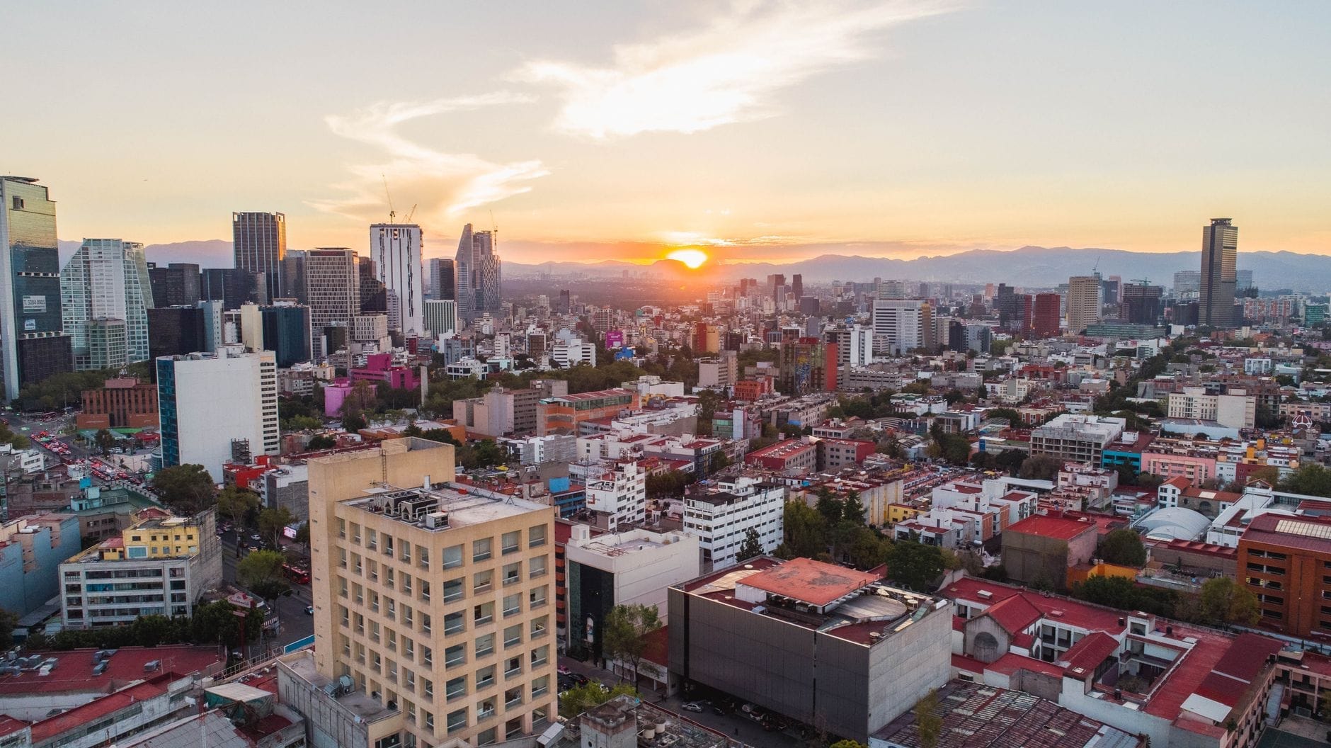 Vista aérea do horizonte da Cidade do México ao pôr do sol com edifícios modernos, áreas residenciais e montanhas ao fundo.