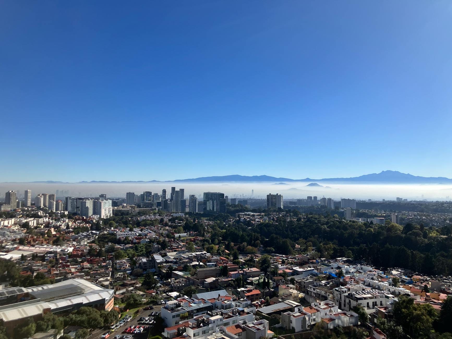 Vista clara do horizonte de Cidade do México com edifícios modernos, bairros e montanhas ao fundo sob um céu azul brilhante e leve névoa.