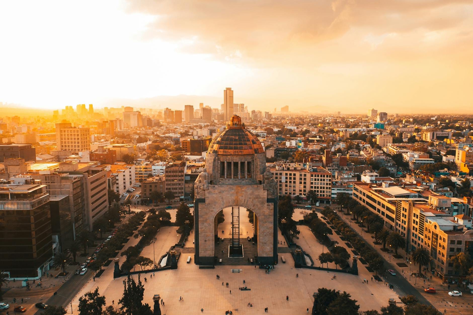 Vista aérea do Monumento à Revolução na Cidade do México ao pôr do sol, cercado por prédios e um céu vibrante.
